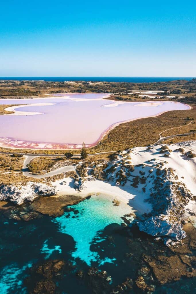 Aerial view of Rottnest Island showcasing the striking pink hues of its salt lake adjacent to the vibrant turquoise of the ocean, with sandy white shores and vegetation in between.