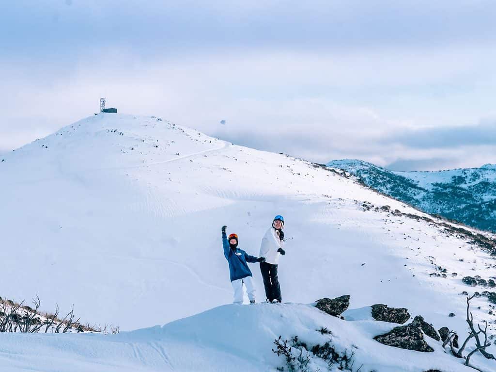 Mt McKay at Falls Creek covered in snow with two women throwing snow in air. 